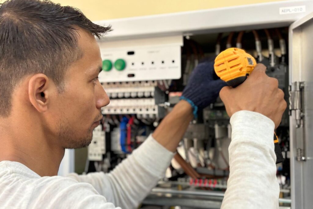 Electrician in work gloves using a cordless drill on an open electrical distribution panel