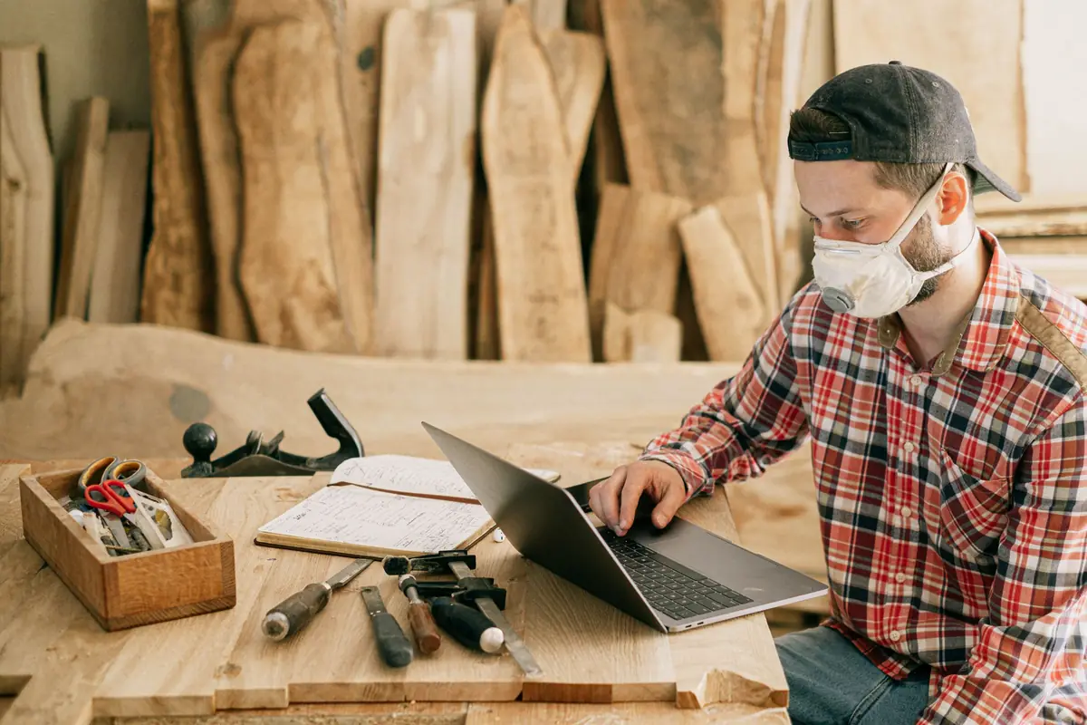 Contractor in a plaid shirt working on a laptop at a workbench in a wood shop