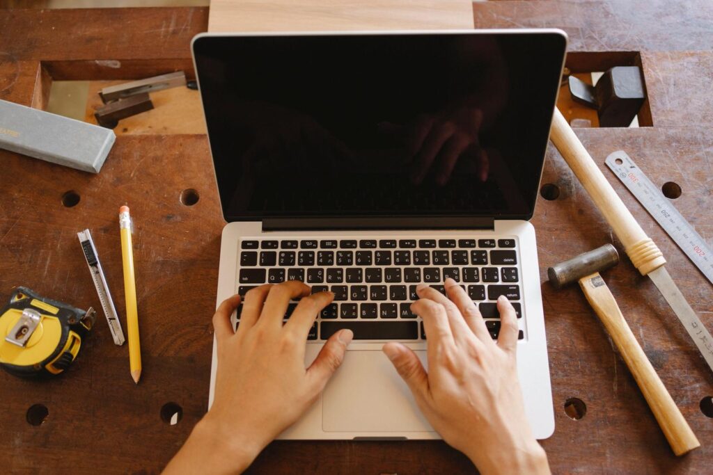 Business owner typing on a laptop on a workbench surrounded by hammers, a tape measure, and hand tools