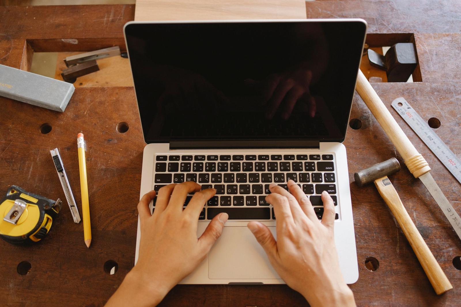 Business owner typing on a laptop on a workbench surrounded by hammers, a tape measure, and hand tools