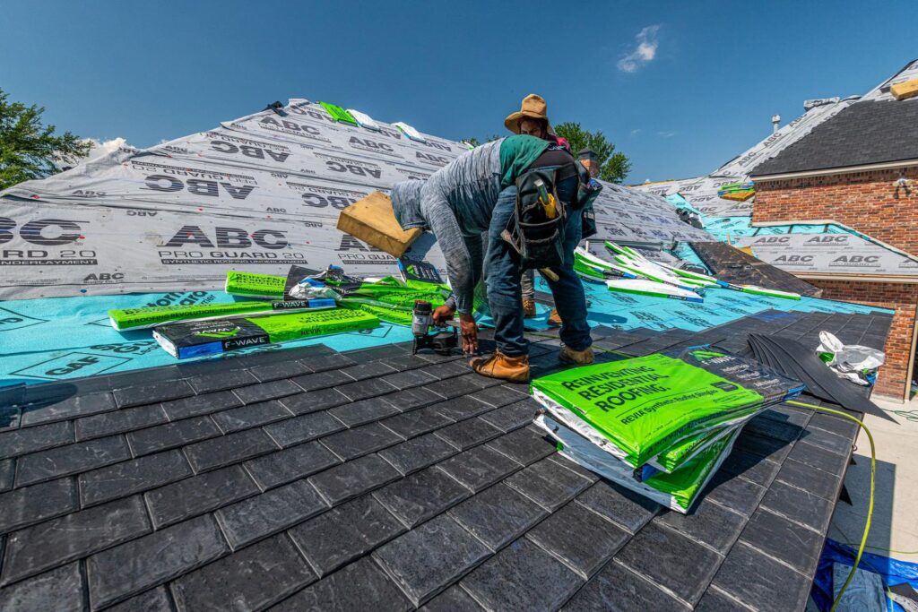 Roofers installing underlayment and shingles on a brick house under a clear blue sky