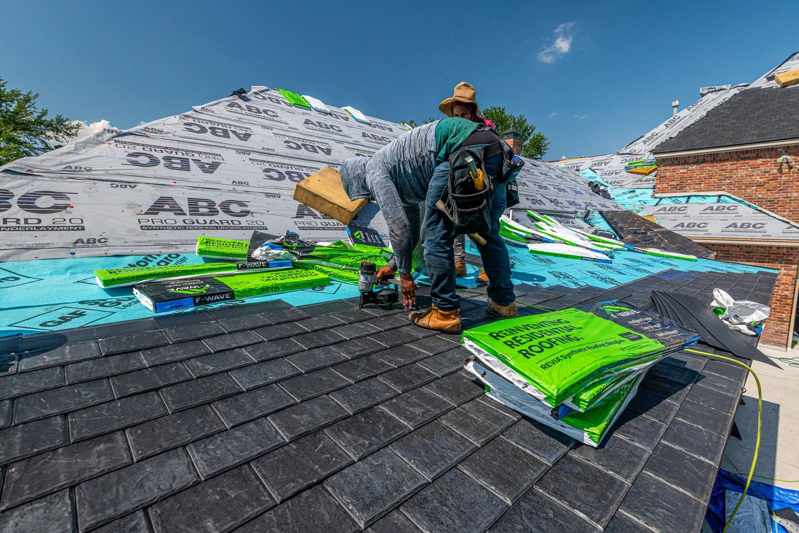Roofers installing underlayment and shingles on a brick house under a clear blue sky