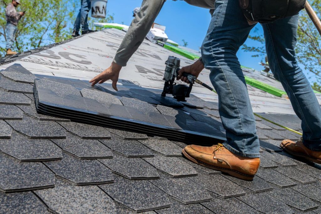 Roofer kneeling on a sloped roof using a nail gun to install dark asphalt shingles