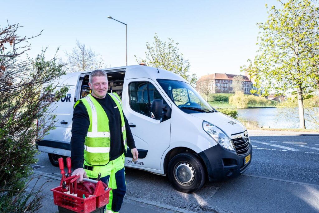 Service technician in a high-visibility vest carrying a tool crate beside a white work van on a residential street