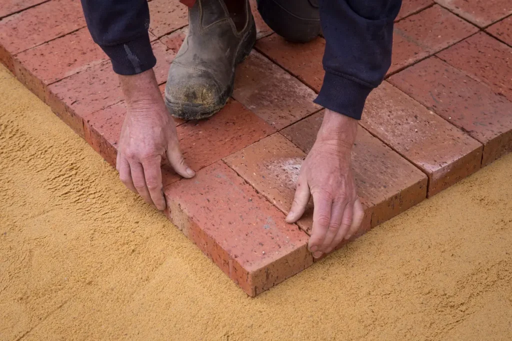 Worker kneeling on sand to set red brick pavers for a new patio